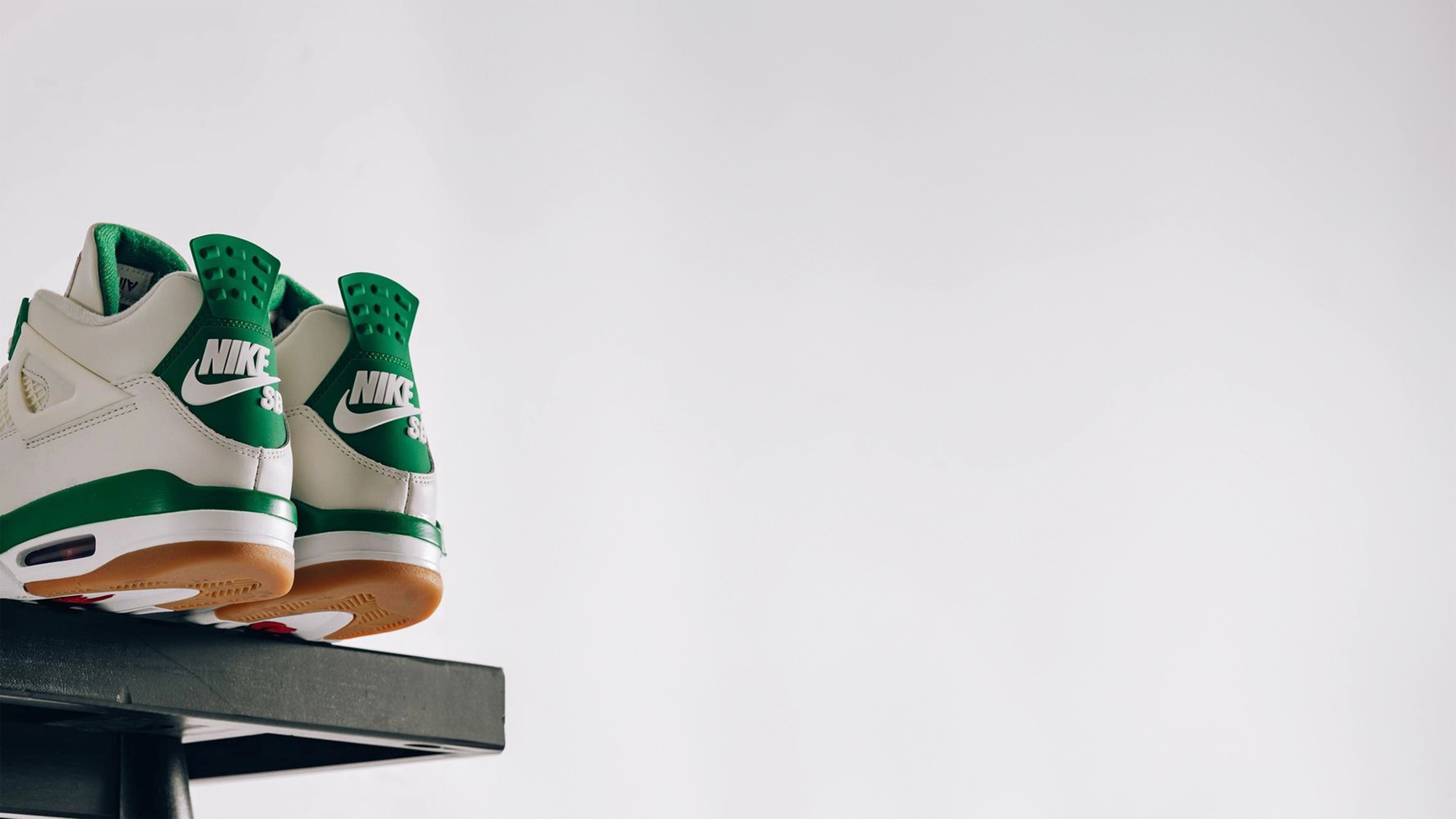 A pair of white and green Nike SB sneakers are displayed on a black platform against a plain white background. The sneakers feature green accents and a gum sole, with the Nike SB logo prominently visible on the heel tabs.